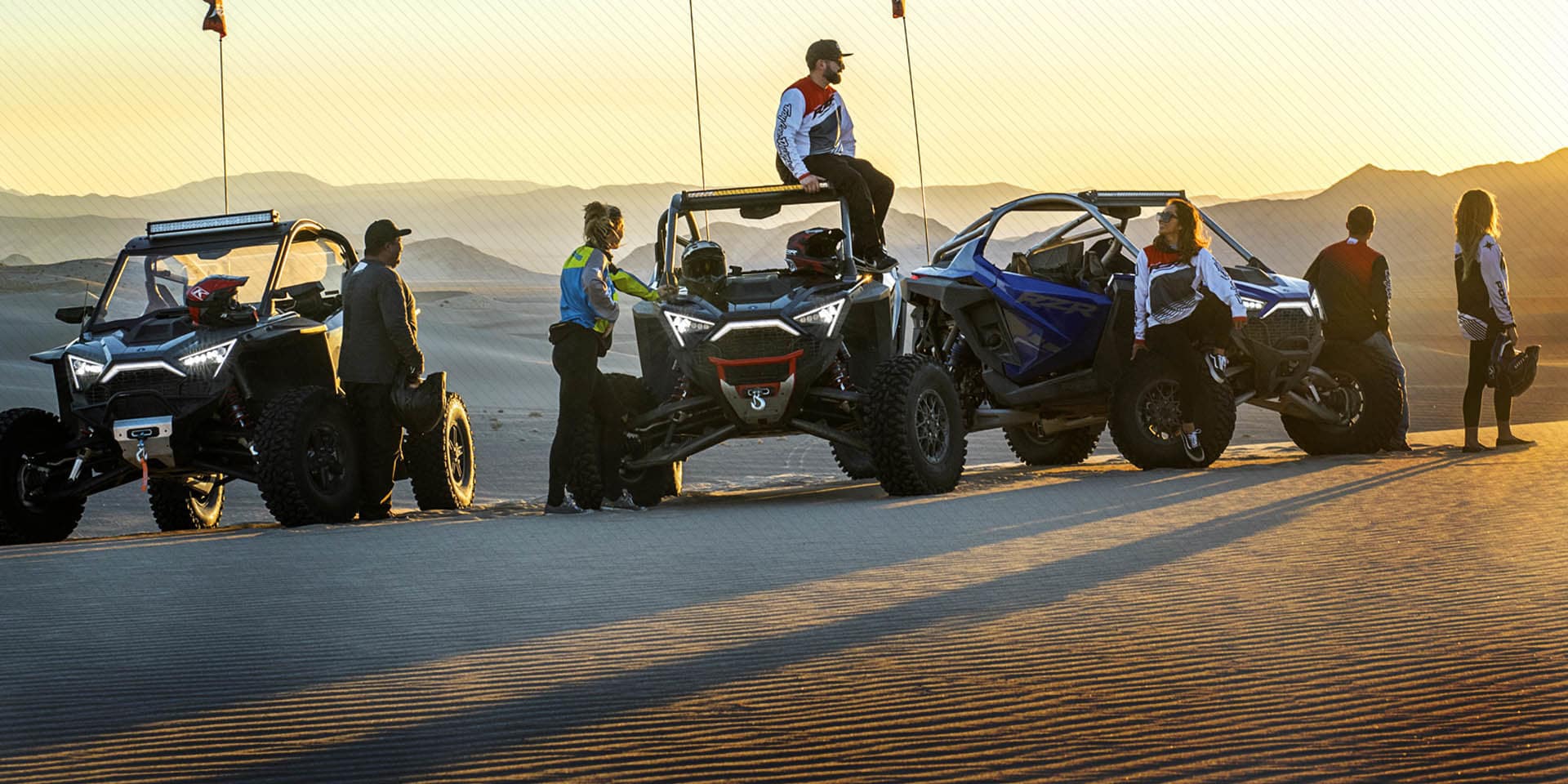 A group of side by side riders gather around their vehicles in a sandy area at sunset.
