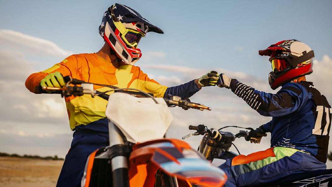 A pair of people in Motorcross gear, are sat on dirt bikes, both giving each other a friendly fist bump. 