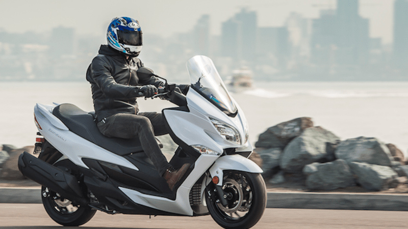 A man on a Suzuki 650 Burgman, that is in white fairing, with a downtown city in the background. 
