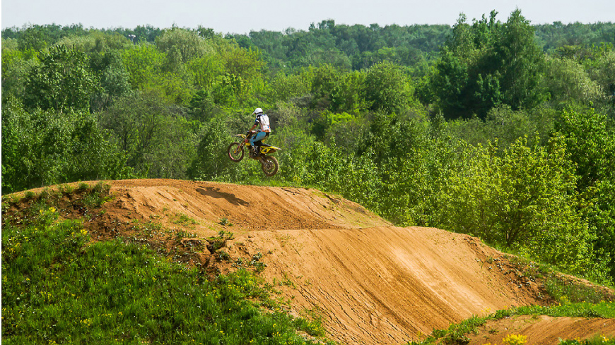 A dirt bike rider on a hilly off-road dirt track surrounded by greenery, jumping through the air over the hill.